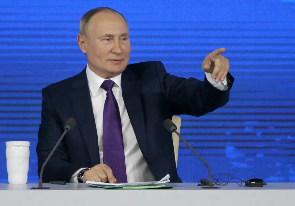 Vladimir Putin points from a table with two microphones and white cup in front of a blue backdrop.
