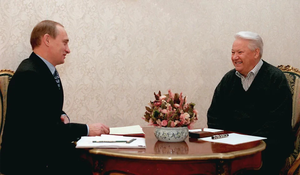 Vladimir Putin and Boris Yeltsin smile while seated at a small round table on which rest flowers, papers and pens.