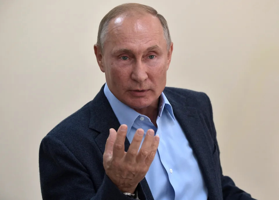 Vladimir Putin, against a beige wall, holds his hand out, gesturing with his fingers pointing upward.