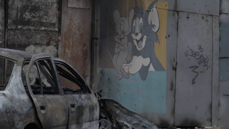 Kuvassa näkyy palanut auto pommituksen jälkeen ja Tom ja Jerry -seinämaalaus.