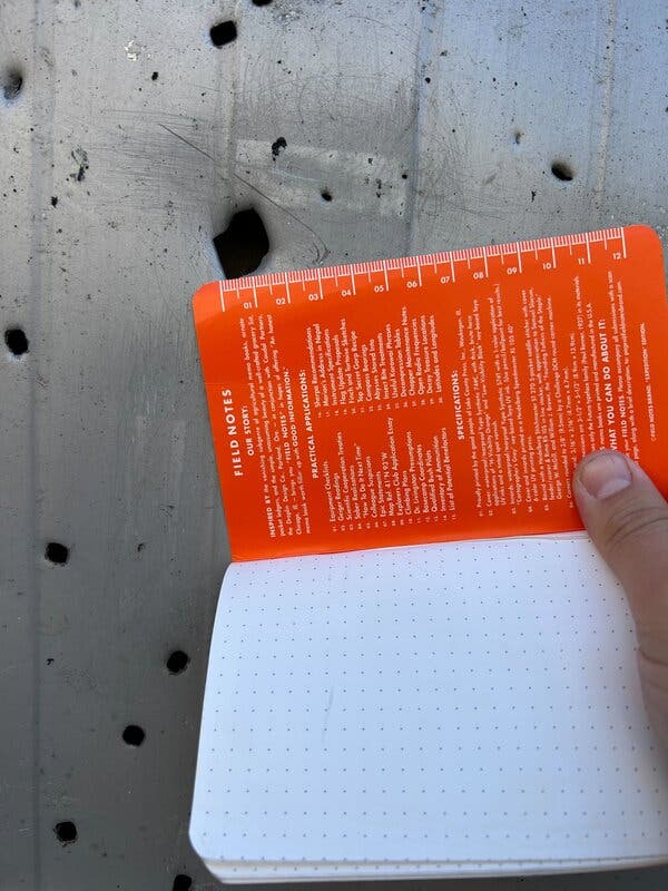 A notebook with a ruler, held next to a hole in a metal facade, showing the hole to be a bit more than one centimeter across.