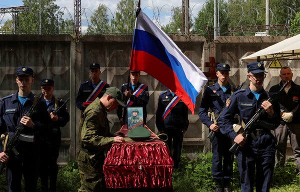 A man in camouflage stands next to a coffin covered in gold-trimmed red cloth and surrounded by soldiers standing at attention. One soldier holds a Russian flag.