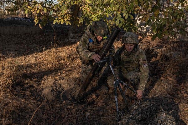 Two soldiers in camouflage with a mortar under a tree.
