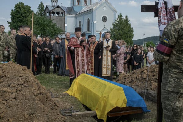 A coffin is covered with a yellow and blue flag. A group of men in religious clothes stands near.