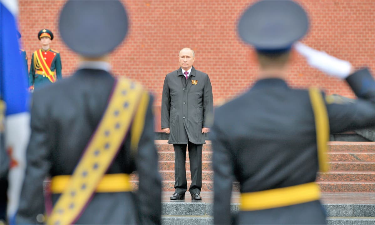 Sotilaat tekevät kunniaa Putinille, joka seisoo harmaassa päällystakissa portailla. Takana näkyy Kremlin punainen muuri.