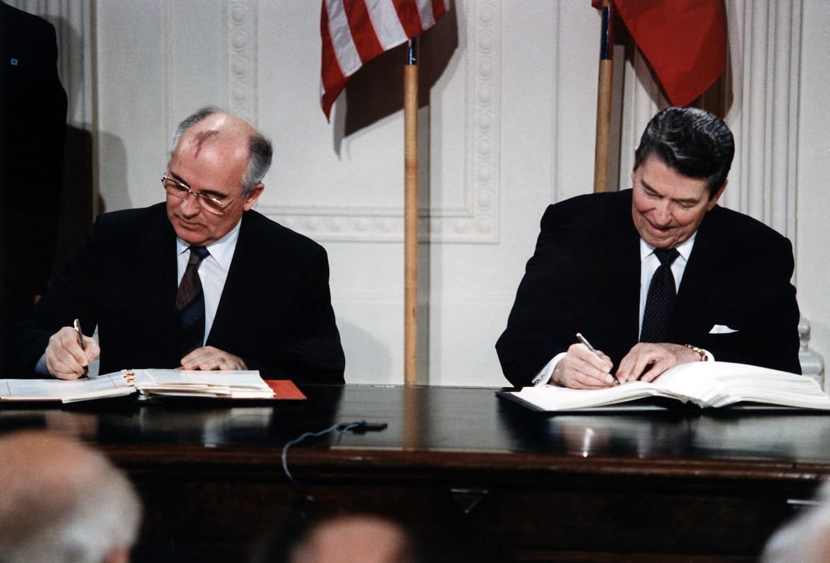 Gorbatšov ja Reagan istuvat pöydän takana ja allekirjoittavat sopimusasiakirjan kappaleita. Miehillä on mustat puvut ja kravatit. Taustalla seinällä ovat maiden liput.