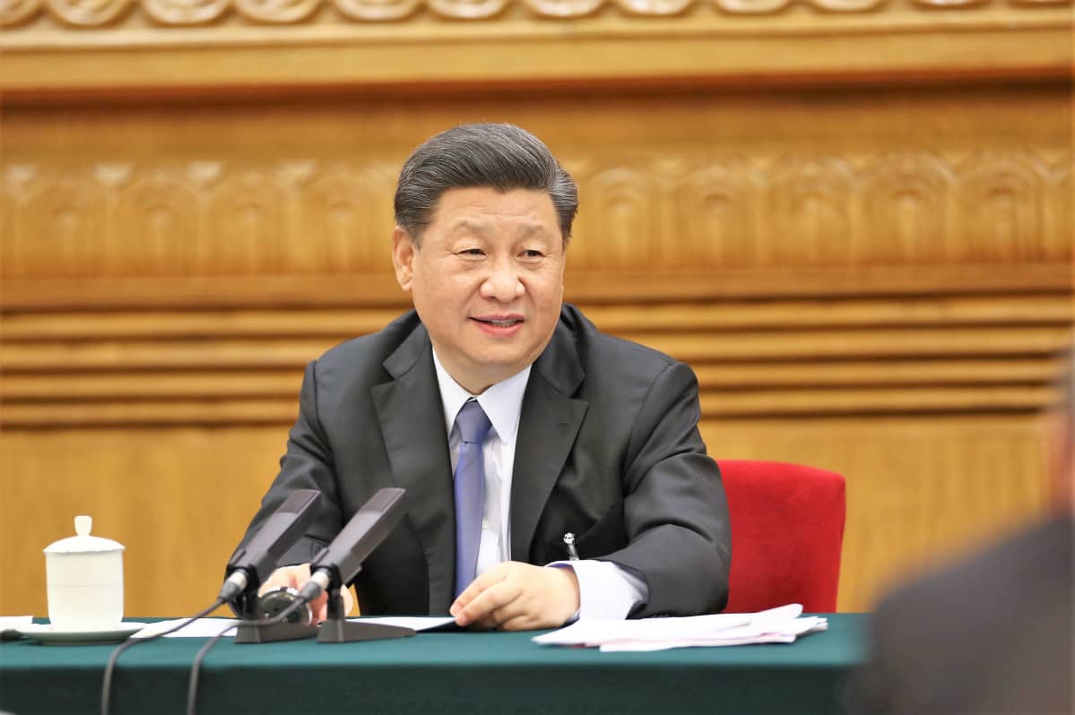 Presidentti Xi Jinping istuu pöydän takana ja puhuu mikrofoniin. Hänellä on edessään papereita. Presidentillä on tummanharmaa puku ja vaalenasininen kravatti.