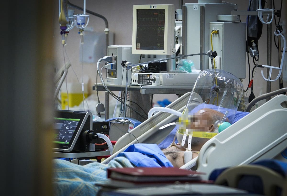 Potilas makaa sairaalavuoteessa hengitysmkoneessa, sängyn ympärillä useita sairaalalaitteita. Kasvot pixelöity.