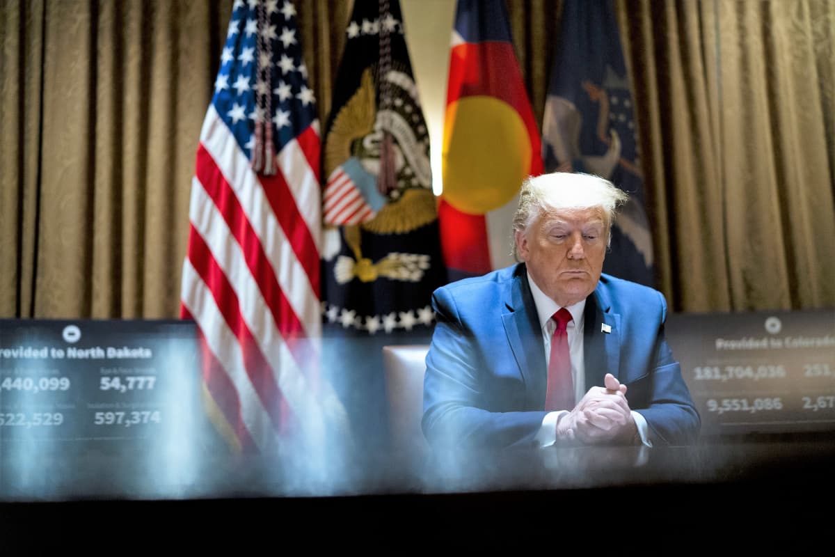 Trump istuu pöydän ääressä ja puristaa käsiään yhteen katsoen alaviistoon. Taustalla näkyy Yhdysvaltain lippu ja muita lippuja. Kuvan vasemmassa reunassa näkyy tietokoneen ruutu, jossa on teksti Pohjois-Dakota ja joitakin lukuja.